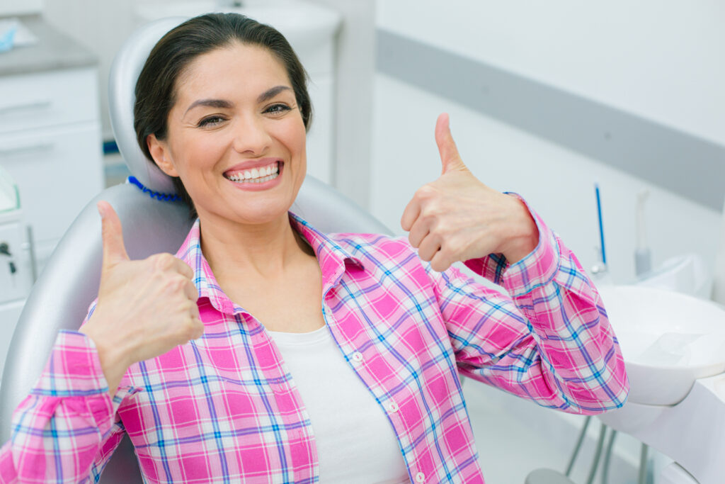 Positive woman revealing her teeth while smiling and showing super gesture with both hands in the dental chair